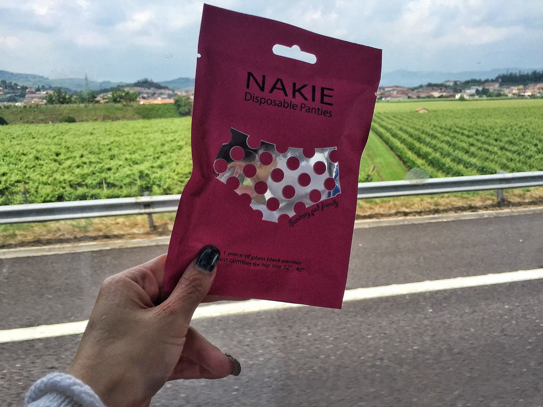มือถือซอง NAKIE ริมถนนที่มีไร่องุ่นและหมู่บ้านบนเนินเขาอิตาลีอยู่ด้านหลัง ทริปเดินต่อได้แม้กระเป๋าไม่มา
